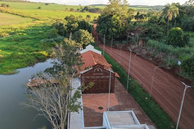 Esta imagem oferece uma perspectiva aérea da Estação Sobradinho e seus arredores, revelando como a construção se integra à paisagem rural de Uberlândia. A foto destaca o telhado de telhas cerâmicas e a fachada lateral de tijolos, situando o edifício entre uma estrada de terra avermelhada, ladeada por postes de iluminação modernos, e um corpo d'água tranquilo à esquerda.