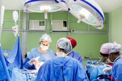 Fotografia em plano médio de uma sala de cirurgia iluminada por dois grandes focos refletores no teto. No centro, uma equipe médica com aventais e gorros azuis, máscaras cirúrgicas brancas e luvas esterilizadas atua concentrada ao redor do paciente, que está coberto por lençóis cirúrgicos azuis. No canto esquerdo, há bolsas de soro penduradas em um suporte metálico; à direita, uma profissional com gorro estampado manuseia instrumentos sobre uma mesa auxiliar. Ao fundo, sob paredes em tom verde-claro, sobressaem-se dois aparelhos de ar-condicionado instalados na estrutura metálica da sala.