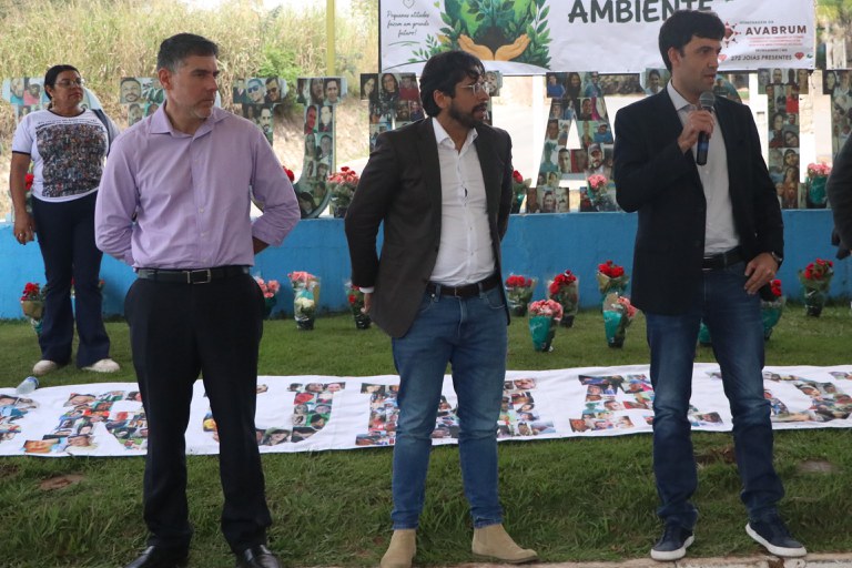 Uma fotografia ao ar livre mostra um grupo de pessoas reunidas em um gramado durante um evento em homenagem às vítimas do rompimento da barragem de Brumadinho. Em primeiro plano, quatro homens estão em pé, lado a lado: da esquerda para a direita, o primeiro veste camisa lilás e calça preta; o segundo veste blazer marrom, camisa branca e calça jeans; o terceiro fala ao microfone vestindo blazer escuro e calça jeans; e o quarto, de terno escuro, está com o corpo levemente inclinado para a frente. Ao fundo, à esquerda, uma mulher vestindo uma camiseta com colagens de fotos observa o grupo, e à extrema direita, parte do corpo de outras duas pessoas é visível. Atrás dos homens, há vasos de flores vermelhas enfileirados no chão e grandes letras decoradas com pequenas fotos de rostos, posicionadas diante de uma mureta azul. Acima deles, destaca-se uma grande faixa branca com uma ilustração do planeta Terra sustentado por mãos e os dizeres "05 de junho - Dia Mundial do MEIO AMBIENTE", acompanhada pelo logotipo da "AVABRUM" (Associação dos Familiares de Vítimas e Atingidos pelo Rompimento da Barragem de Brumadinho).