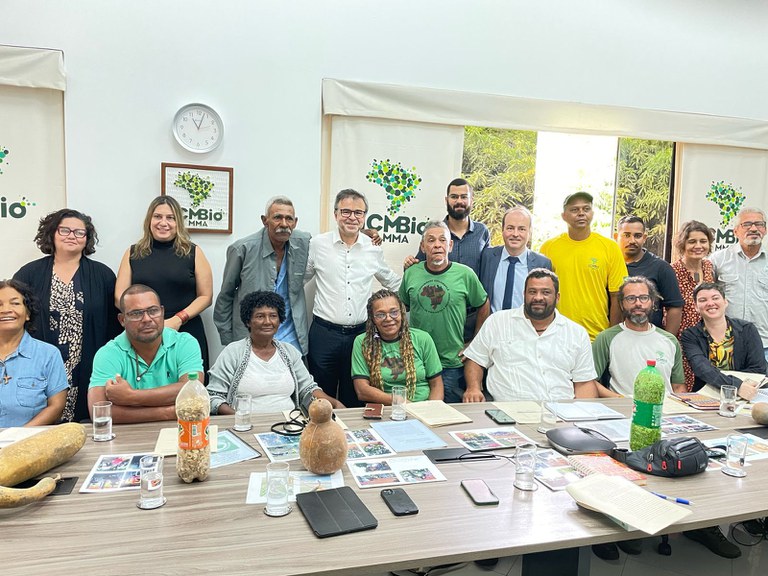 a foto mostra um grupo diverso de vinte pessoas sorrindo para a câmera em uma sala iluminada, reunidas em torno de uma mesa com objetos variados e um banner com o logotipo "ICMBio" ao fundo. Registro de uma reunião.