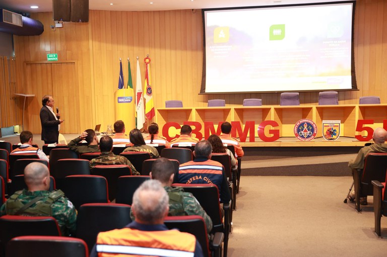 Fotografia do auditório da prefeitura de Uberlândia, com pessoas sentadas na platéia, um homem de terno em pé, falando ao microfone e olhando em direção ao telão, ao fundo.