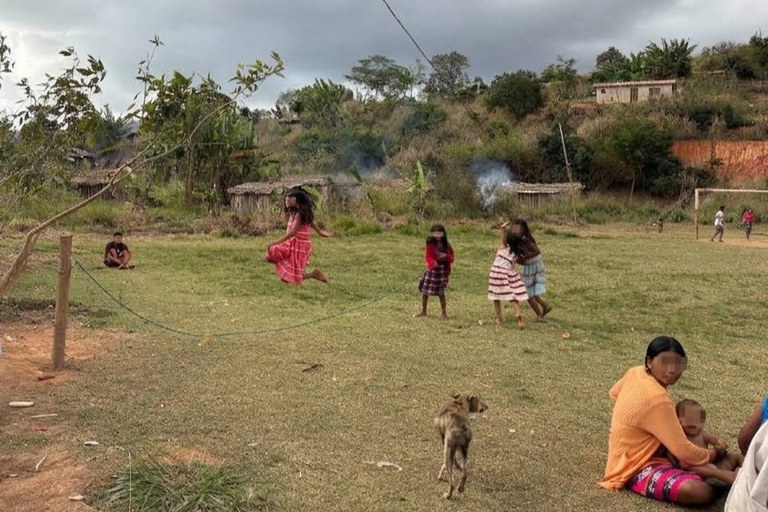 Fotografia ao ar livre, em uma área rural e descampada com gramado baixo e solo de terra batida, mostrando crianças e adultos indígenas no povoado. No centro, uma menina de vestido rosa salta sobre um elástico esticado na horizontal preso a postes de madeira, enquanto outras três crianças assistem por perto e um cachorro magro caminha na grama. No canto inferior direito, uma mulher sentada no chão segura um bebê no colo, acompanhada por outra pessoa parcialmente visível. Ao fundo, veem-se morros com vegetação rasteira, pequenas casas simples de madeira e palha, traves de um campo de futebol improvisado e um céu nublado.