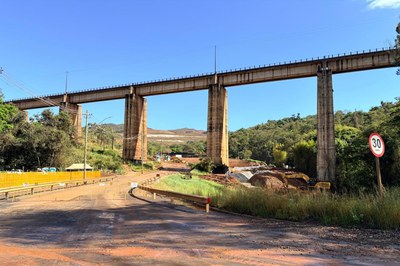 Fotografia em ângulo intermediário de um elevado ferroviário de concreto sustentado por altos pilares, atravessando uma área de obras e mineração sob céu azul limpo. Abaixo da estrutura, uma estrada de terra avermelhada e pavimentação com poeira passa por barreiras amarelas de proteção, defensas metálicas e uma placa redonda de trânsito indicando limite de velocidade de 30 km/h no canto direito. Ao fundo, entre a vegetação dos morros, observa-se o maquinário amarelo em operação e encostas com terra exposta.