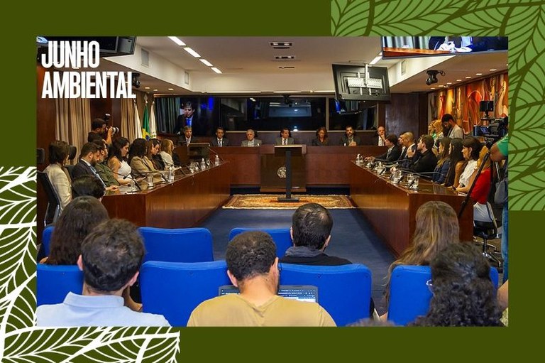 Foto mostra auditório onde foi realizado o evento de assinatura do acordo com duas mesas nas laterais com diversas pessoas entre homens e mulheres sentadas com roupas formais, ao fundo, formando um U com as mesas laterais, outra mesa com sete pessoas entre homens e mulheres também com roupas formais, as mesas possuem microfones e copos de água, No canto inferior, em primeiro plano, pessoas acompanham o evento em cadeiras azuis