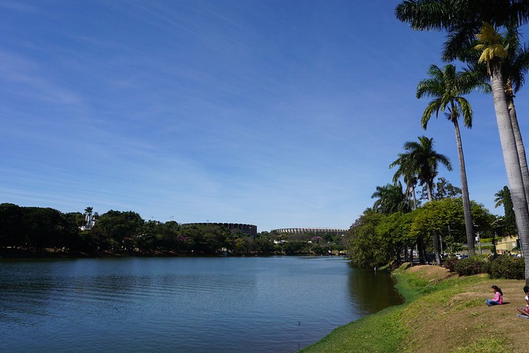 Vista diurna e panorâmica da Lagoa da Pampulha em Belo Horizonte. A lagoa de água calma preenche o centro, com margens arborizadas e palmeiras altas à direita. Ao fundo, visíveis acima da vegetação, estão o Mineirão (Estádio Governador Magalhães Pinto) e, à esquerda, o Mineirinho (Ginásio Jornalista Felippe Drummond).