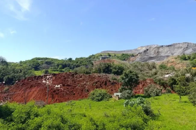Vista panorâmica de uma área rural com vegetação e terra exposta. Ao fundo, uma grande colina com pedras de mineração visíveis no topo.