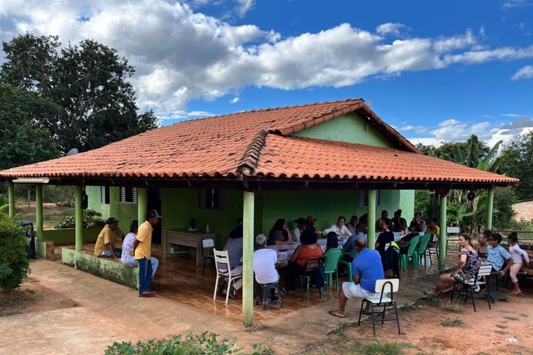 Uma reunião comunitária acontece na varanda coberta de uma casa simples com paredes verdes e telhado de telhas cerâmicas vermelhas. Cerca de quinze pessoas estão reunidas em torno de uma mesa e dispostas em cadeiras espalhadas sob a varanda e no quintal de terra batida ao redor. O local é cercado por vegetação densa e árvores ao fundo, sob um céu azul ensolarado e com nuvens brancas bem definidas.