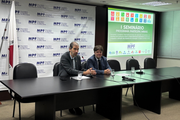 A imagem mostra um ambiente de conferência ou seminário, possivelmente no Ministério Público Federal (MPF) em Minas Gerais, como indicado pelos logotipos e textos no fundo. Dois homens estão sentados em uma mesa preta na frente de uma tela de projeção. O homem à esquerda tem barba e veste um terno escuro e gravata clara, enquanto o homem à direita veste um terno azul-marinho e gravata escura. A mesa tem microfones e papéis na frente deles. Na tela de projeção, em destaque, está escrito "I SEMINÁRIO Programa Participa Minas" e, abaixo, uma descrição sobre "Fomento a projetos intersetoriais de extensão universitária em interface com a pesquisa visando ao desenvolvimento sustentável e prevenção, mitigação e correção dos impactos da mineração no Estado de Minas Gerais". No topo da tela, são visíveis os ícones dos Objetivos de Desenvolvimento Sustentável (ODS) da ONU.