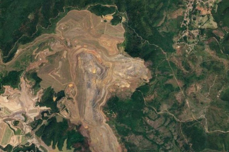 Vista de satélite de uma grande área de mineração a céu aberto, destacando o solo escavado em tons de marrom cercado por densa vegetação verde.