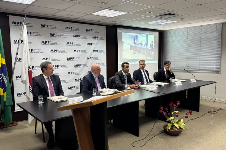 Foto mostra mesa do evento com os palestrantes, por trás das pessoas a bandeira do Brasil e outra bandeira, um banner do MPF e um telão com a transmissão do evento