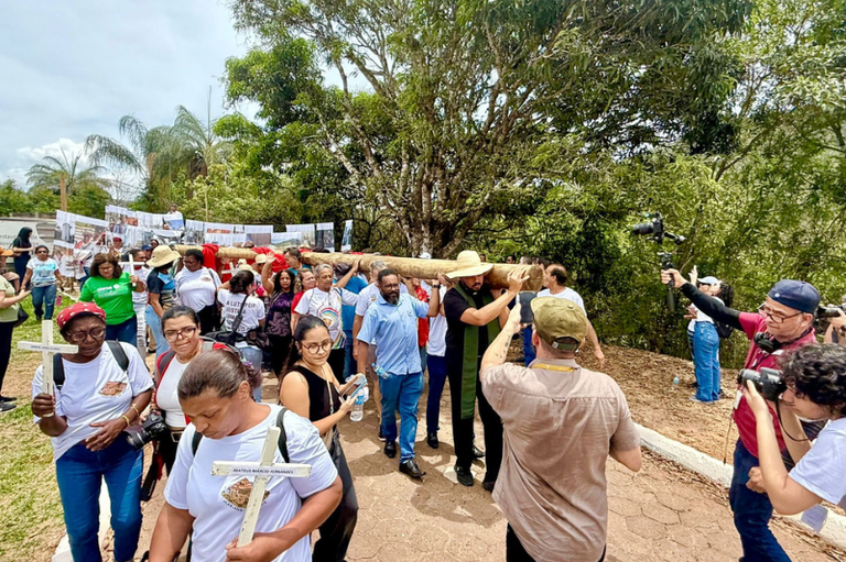 Cortejo com dezenas de pessoas, algumas carregando uma grande cruz de madeira e outras segurando pequenas cruzes brancas, em ato. Ao fundo, faixa com fotos e árvores.