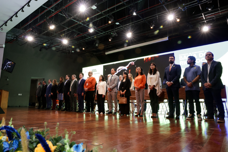 Foto mostra cerca de 20 pessoas em palco de auditório, de pé lado a lado. Ao fundo, por trás das pessoas, há um painel grande onde se lê Bacia do Rio Doce