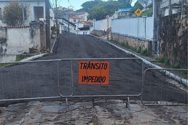 Foto mostra parte da rua asfaltada e parte com o calçamento original. Grades delimitam o local e um placa laranja informa: trânsito impedido
