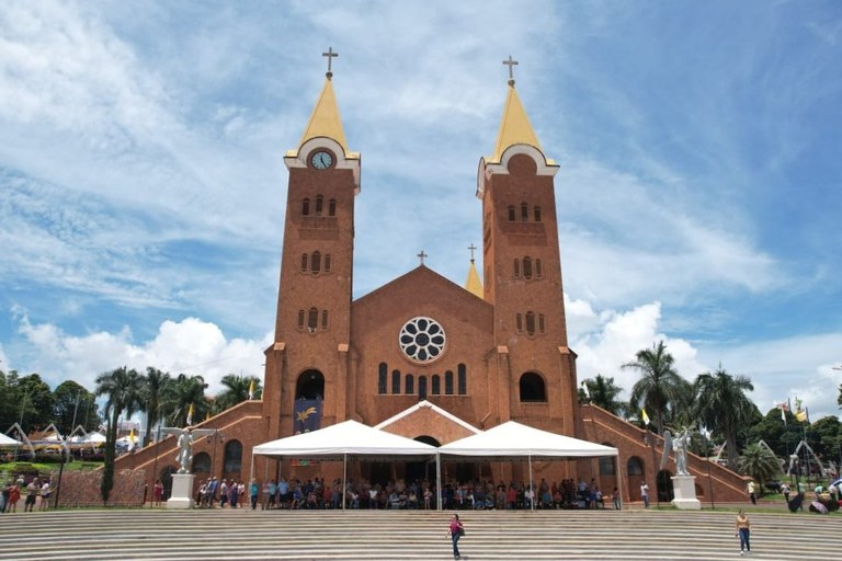 Esta imagem apresenta a imponente fachada do Santuário de Nossa Senhora da Abadia, localizado em Romaria, Minas Gerais. A estrutura é construída em tijolos aparentes de tom terroso, destacando-se por suas duas torres simétricas com agulhas amarelas vibrantes e uma rosácea central acima da entrada. O cenário exibe um dia ensolarado com céu azul e nuvens esparsas, enquanto uma multidão de fiéis se reúne à frente do templo, protegida por tendas brancas montadas sobre uma ampla escadaria de pedra. A composição é emoldurada por palmeiras e elementos religiosos, como estátuas de anjos e bandeiras, transmitindo uma atmosfera de devoção e importância cultural para a região do Triângulo Mineiro.