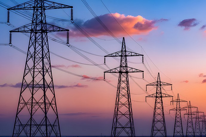 Esta imagem apresenta uma fileira de torres de transmissão de energia elétrica que se estende em direção ao horizonte, capturada durante o entardecer ou amanhecer. As estruturas metálicas imponentes aparecem como silhuetas escuras e detalhadas contra um céu vibrante, que exibe um gradiente de tons suaves de azul, roxo e rosa-alaranjado. Algumas nuvens esparsas, iluminadas pela luz solar indireta, flutuam sobre as linhas de alta tensão, conferindo um aspecto quase artístico e sereno a um cenário tipicamente industrial e de infraestrutura.