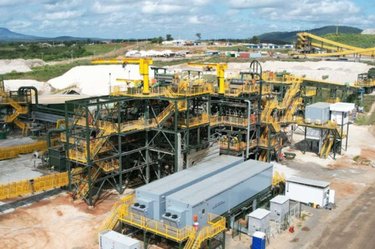 Complexo industrial com maquinário e grandes tubulações, em meio a montes de minério branco. A imagem representa a exploração de lítio em Minas Gerais.