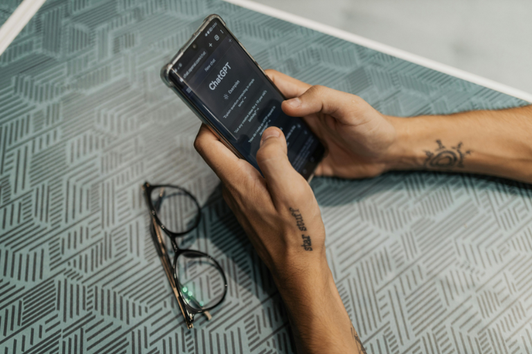 Foto mostra detalhe de braços de homem com algumas tatuagens apoiados sobre uma mesa com um telefone celular na mão. No aparelho, vê-se uma tela do ChatGPT. Sobre a mesa há um par de óculos de grau.