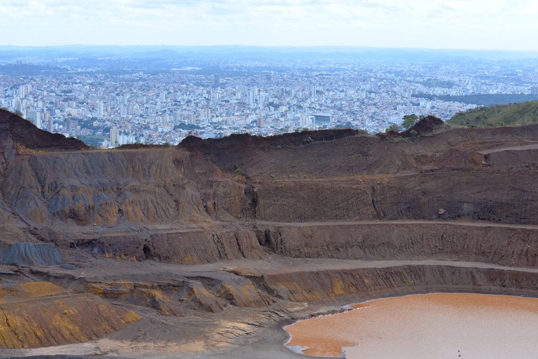 Foto mostra, em primeiro plano, grande barranco desmatado pela mineração e, ao fundo e a distância, vista aérea de uma cidade