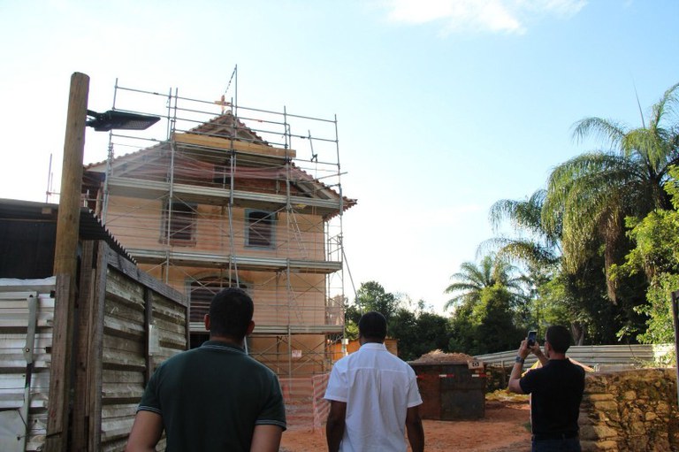 Foto mostra uma casa de madeira em construção com andaimes na frente e três pessoas olhando para ela