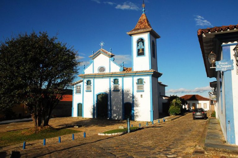 Igreja colonial com fachada branca, detalhes em azul e torre sineira, situada em uma praça com calçamento de paralelepípedos sob céu azul.