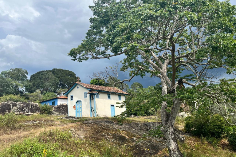 Foto mostra uma igreja antiga, branca e azul, em meio à uma paisagem típica do cerrado mineiro.