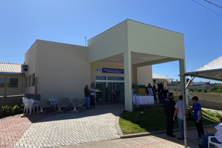 Fachada da Unidade Básica de Saúde Edson Paulo de Souza sob céu claro. Pessoas circulam na entrada do prédio moderno, que possui rampa de acessibilidade e áreas gramadas.