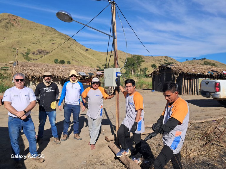 Grupo de seis homens posa em frente a poste com luminária e caixa elétrica em comunidade indígena. Ao fundo, veem-se habitações simples de palha e madeira sob céu ensolarado e montanhas ao fundo.