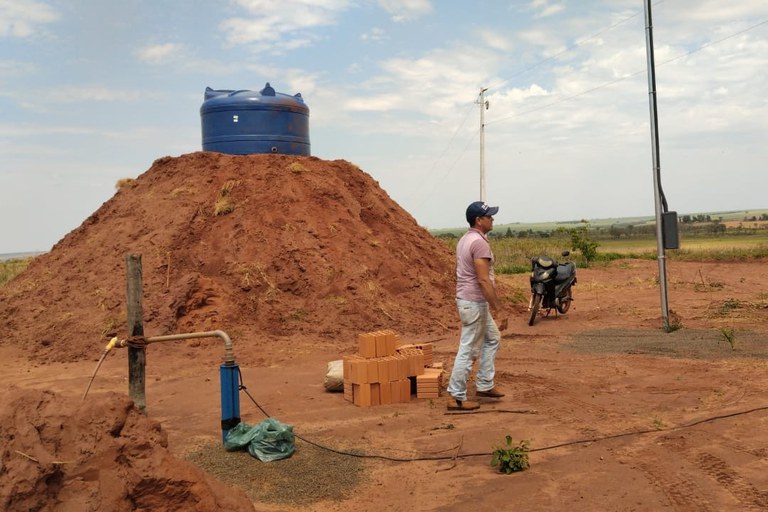 Fotografia colorida em plano aberto de um poço artesiano instalado em uma área rural indígena. Há um morro de barro com uma caixa d´água azul em cima. Ao redor, há um homem andando, uma moto ao fundo e o chão de terra batida se mistura com vegetação rasteira e arbustos. No plano de fundo, visível do centro para a direita, há uma área de mata densa. A luz do dia sugere que o trabalho está em andamento.