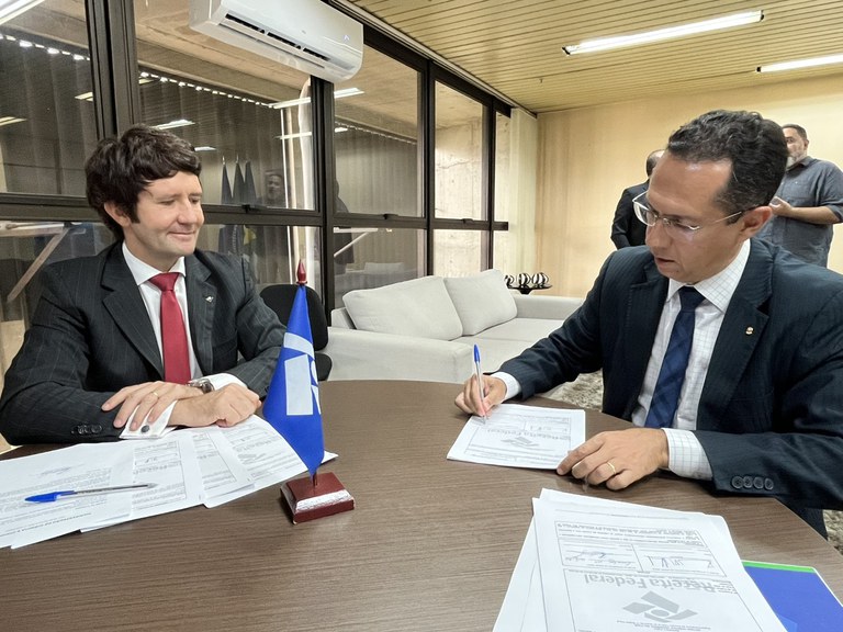Em uma sala de reunião, sobre uma mesa de madeira escura redonda, os dois representantes do MPF e do RF assinam documentos. O procurador-chefe Ricardo Pael está à direita, de terno azul escuro, e se debruça sobre o papel, com uma caneta na mão, assinado o documento. Do lado esquerdo, está o delegado chefe da RF, Raimundo Sari, que sorri e contempla o colega assinando o ato de doação.