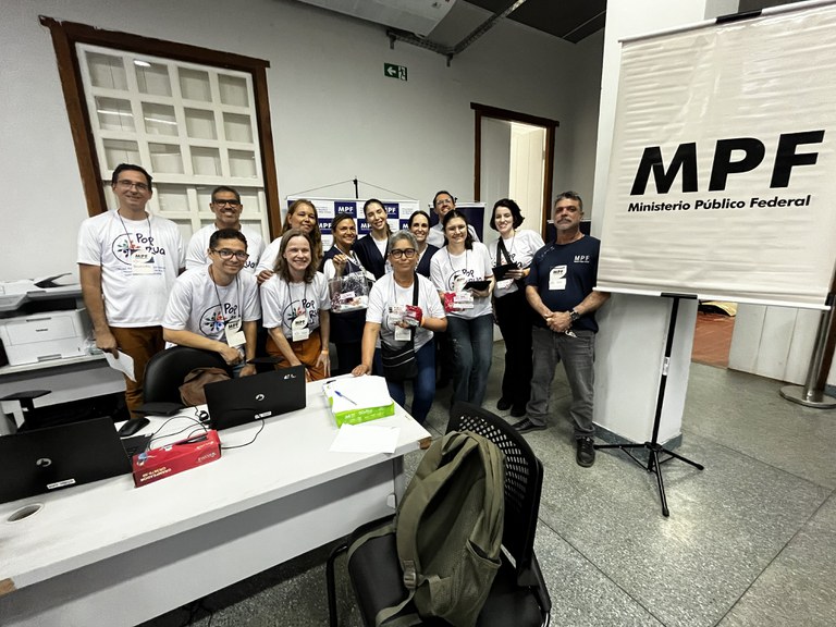 Foto em ambiente interno reúne uma equipe de mais de dez pessoas sorrindo atrás e ao lado de uma mesa de escritório com notebooks e papéis.
A maioria veste camisetas brancas com a estampa "Pop Rua" e crachás, enquanto à direita há um banner retrátil branco com a sigla "MPF" e a inscrição "Ministério Público Federal".