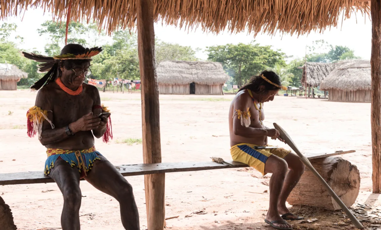 A imagem mostra dois homens indígenas sentados em um banco sob um teto de palha em uma aldeia. À esquerda, um homem com cocar e trajes tradicionais foca sua atenção mexendo em um smartphone. À direita, o outro homem, vestindo bermuda e adereços, trabalha esculpindo uma longa peça de madeira. Ao fundo, o pátio de terra batida revela outras ocas tradicionais e vegetação sob a luz forte do dia.