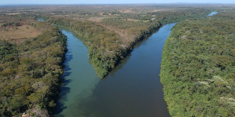Foto mostra bifurcação do Rio Cuiabá em dia claro e ensolarado, com cada uma das vertentes apresentando uma coloração da água: do lado esquerdo, a água é verde, da outra, marrom escuro. Entre os dois cursos do rio e nas laterais, densa floresta com algumas falhas provocadas por atividade humana.
