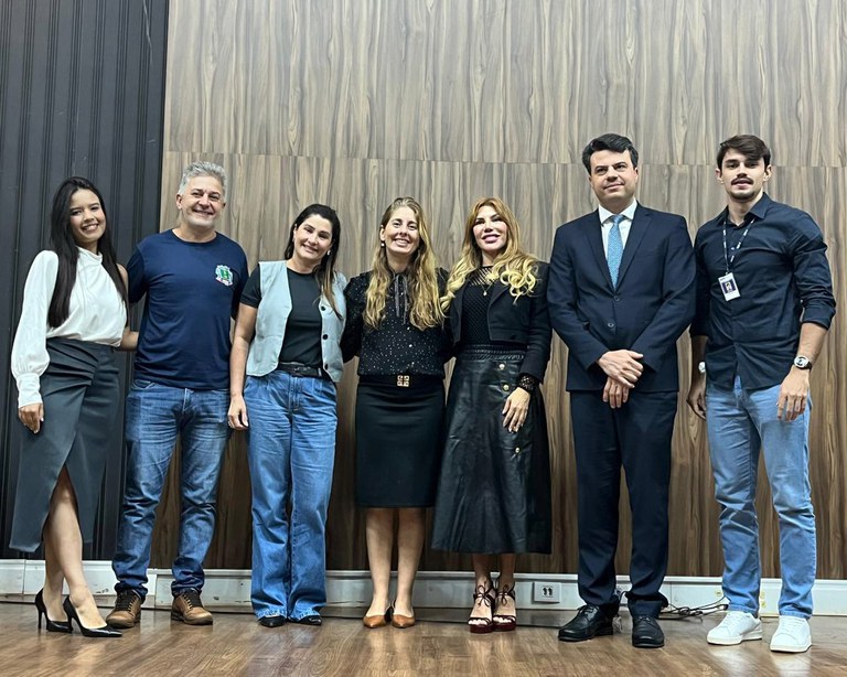 Sete pessoas posam lado a lado em um ambiente com piso de madeira e parede de fundo amadeirada. À esquerda, três pessoas vestem trajes casuais, seguidas por três mulheres com roupas sociais escuras (duas de saia e uma de calça) e, na ponta direita, um homem de terno azul-marinho ao lado de outro jovem de camisa preta e calça jeans.