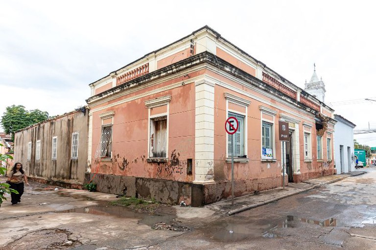 Casarão histórico de esquina em tom rosa-salmão com detalhes brancos, exibindo placa do IPHAN e sinalização de trânsito na calçada.