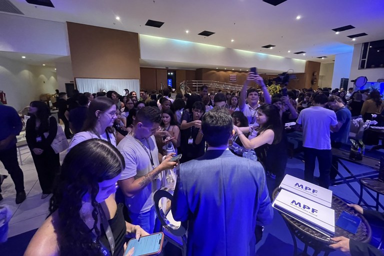 Uma fotografia em plano médio, capturada de um ângulo ligeiramente elevado, mostra um grande grupo de pessoas reunidas no saguão de um evento acadêmico ou corporativo com iluminação em tons de azul e roxo. No centro do primeiro plano, um homem de terno cinza está de costas para a câmera, cercado por jovens que olham em sua direção, muitos deles segurando celulares para tirar fotos ou gravar vídeos. À direita, sobre uma pequena mesa redonda, destacam-se duas pastas ou caixas brancas com o logotipo "MPF" (Ministério Público Federal) em letras azuis. Ao fundo, dezenas de outros participantes conversam e se movimentam pelo amplo espaço do auditório, que possui paredes em tons de bege e marrom.