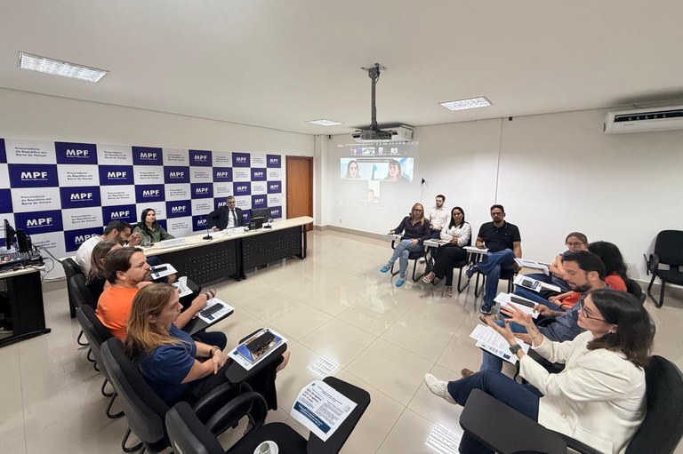 A imagem mostra uma reunião presencial realizada em uma sala de conferências da Procuradoria da República (MPF) em Barra do Garças, Mato Grosso, conforme indicado pelas inscrições no painel ao fundo. Cerca de quinze pessoas estão dispostas em um formato semicircular utilizando cadeiras universitárias pretas com pranchetas acopladas, enquanto um homem de terno e uma mulher com jaqueta verde conduzem os trabalhos sentados atrás de uma mesa principal ao fundo à esquerda. Os participantes, vestidos com roupas casuais e de negócios, prestam atenção a uma mulher de blazer branco em primeiro plano à direita, que gesticula ativamente enquanto fala. Ao fundo, um projetor exibe uma videoconferência na parede branca com duas participantes remotas, indicando um modelo de reunião híbrido, tudo sob a iluminação clara de um teto com luminárias embutidas e piso de porcelanato claro.