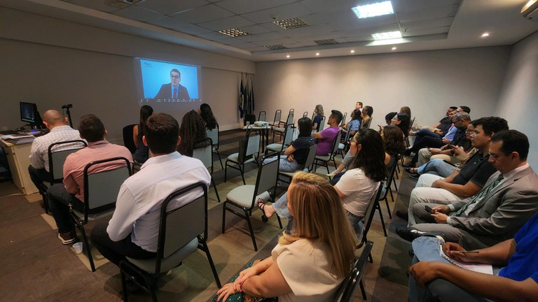 foto de uma sala de treinamento com pessoas sentadas de costas olhando para uma tela projetada na parede onde um homem dá uma palestra