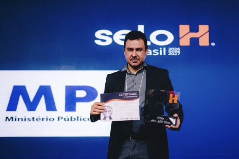 Coordenador de Administração da PR/MT posa para foto. Ele está em pé, segurando o certificado em uma mão e o prêmio em outra. Ele veste uma calça jeans, cinto preto, camisa cinza riscada e blazer preto. No fundo, uma tela azul. Do lado esquerdo, a logo do MPF