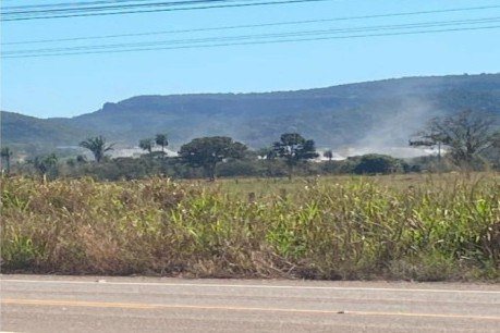 Uma imagem horizontal em plano médio mostra uma paisagem rural vista a partir de uma estrada em um dia ensolarado. No primeiro plano, na borda inferior, há um trecho de asfalto cinza com linhas amarelas pintadas. Logo atrás da estrada, uma faixa de vegetação rasteira e densa, composta por capim alto de tons verdes e secos, estende-se horizontalmente. No plano médio, uma área de pasto plano abriga árvores esparsas e uma estrutura industrial de cor clara, parcialmente encoberta por uma nuvem de poeira ou fumaça branca que se dispersa para a direita. Ao fundo, uma imponente cadeia de montanhas ou chapadas de relevo plano e coloração azulada recorta o horizonte sob um céu azul claro e limpo, cortado horizontalmente por linhas de alta tensão na parte superior da imagem.