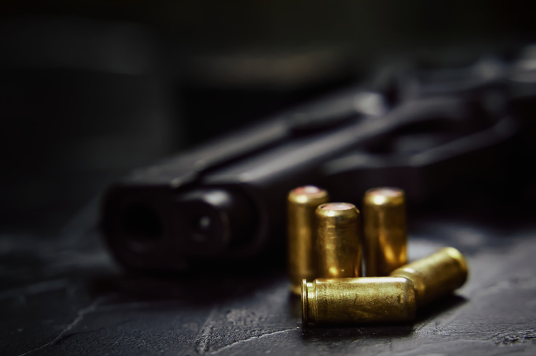 foto com tom escuro mostra arma de fogo preta sobre uma mesa. Ao lado, há munições douradas.
