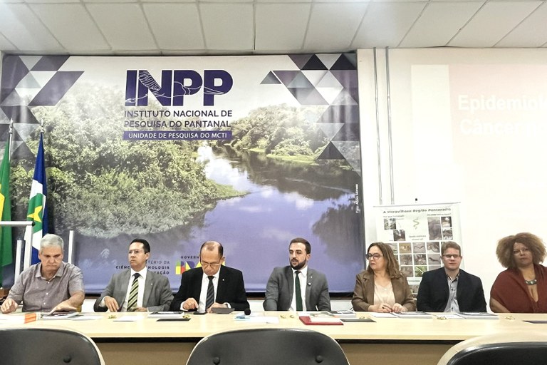 A imagem mostra uma mesa de autoridades composta por sete pessoas (cinco homens e duas mulheres) sentadas lado a lado durante um evento ou reunião institucional. Ao fundo, destaca-se um grande painel com o logotipo do INPP (Instituto Nacional de Pesquisa do Pantanal), uma unidade de pesquisa do MCTI, ilustrado com a foto de um rio sinuoso cercado por vegetação densa e, no canto esquerdo, as bandeiras do Brasil e do estado de Mato Grosso. Os integrantes da mesa estão vestidos formalmente: os homens vestem paletós ou camisas sociais, e as mulheres usam blazers e trajes casuais-chiques, com papéis, pastas e garrafas de água espalhados sobre a mesa clara à sua frente. Do lado direito do painel principal, há um cartaz menor com o título "A Maravilhosa Região Pantaneira" exibindo pequenas fotos da fauna local, sob uma projeção digital parcialmente visível na parede branca.