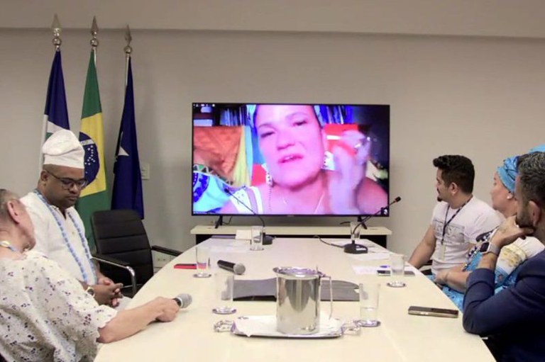 Foto mostra mesa de debate do podcast com cinco pessoas sentadas ao redor olhando para um telão enquanto outra participante fala