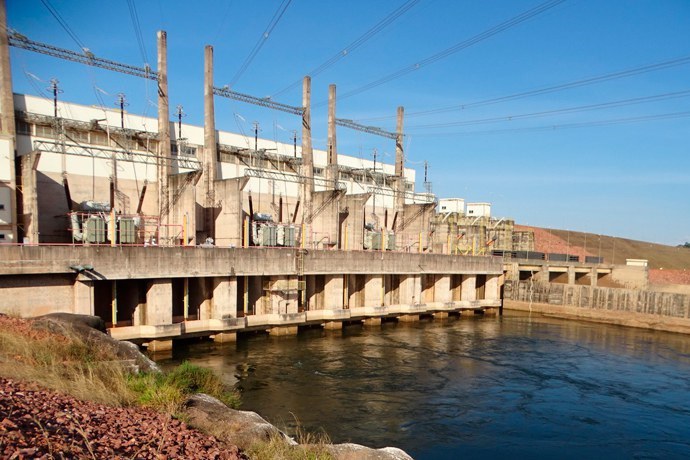 Fotografia da Usina Hidrelétrica Colíder no Rio Teles Pires, em Mato Grosso, mostrando a barragem de concreto e o vertudouro no centro. A estrutura divide a paisagem: de um lado, a água calma e verde-azulada do reservatório; do outro, o rio continua o curso, com o leito parcialmente seco e áreas de terra batida visíveis. A usina está cercada por uma densa vegetação verde de mata atlântica nas margens, sob um céu claro com poucas nuvens.