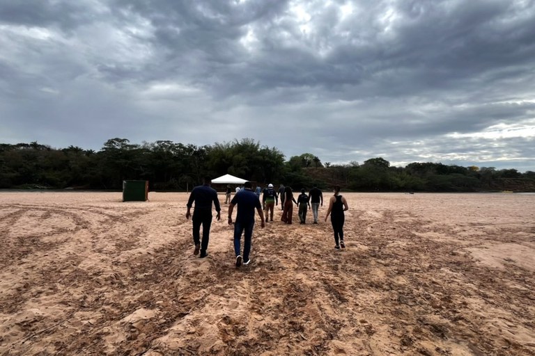 Grupo de pessoas caminhando de costas por uma ampla faixa de areia sob um céu nublado, em direção a uma tenda branca montada próxima a árvores ao fundo.