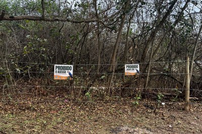 Cerca de arame em frente a uma vegetação densa e seca, com duas placas fixadas: uma à esquerda dizendo "Proibido o Acesso" e outra à direita onde se lê "Proibido Jogar Lixo".
