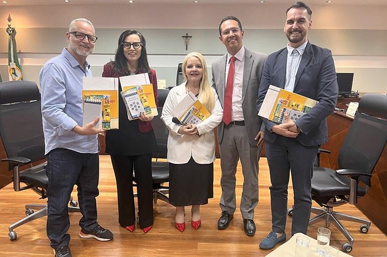 Cinco pessoas posam sorrindo para a foto em um auditório institucional. Três homens e duas mulheres estão em pé, segurando publicações coloridas.