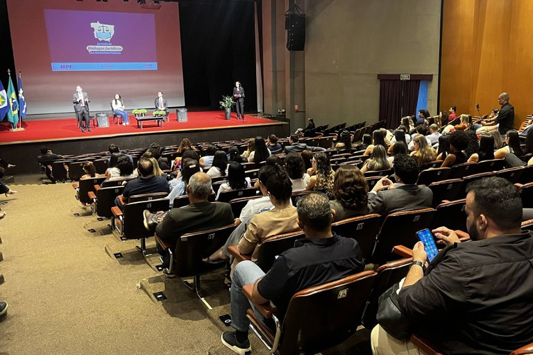 A imagem mostra um auditório amplo visto do fundo da plateia durante a "Jornada de Diálogos Jurídicos dos Ministérios Públicos". Em segundo plano, o palco iluminado possui um telão ao centro exibindo a logomarca do evento e as siglas dos órgãos realizadores (MPF, MPT e MPMT), acompanhado por três palestrantes sentados e um orador em pé à esquerda falando ao microfone. Em primeiro plano e ao longo da plateia, diversas pessoas aparecem sentadas nas poltronas escuras assistindo à apresentação, enquanto algumas interagem discretamente com celulares.