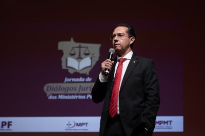 A imagem mostra um homem de pele clara, cabelos curtos escuros e óculos de grau, vestindo terno preto, camisa social branca e gravata vermelha, em um enquadramento de meio-corpo ligeiramente em ângulo baixo. Ele está em pé, segurando um microfone com a mão direita na altura do peito como se estivesse discursando, enquanto mantém a mão esquerda no bolso da calça. Ao fundo, ligeiramente desfocado, vê-se o painel do evento "Jornada de Diálogos Jurídicos dos Ministérios Públicos", com um logotipo contendo uma balança sobre um livro e as siglas do MPF, MPT e MPMT na parte inferior.