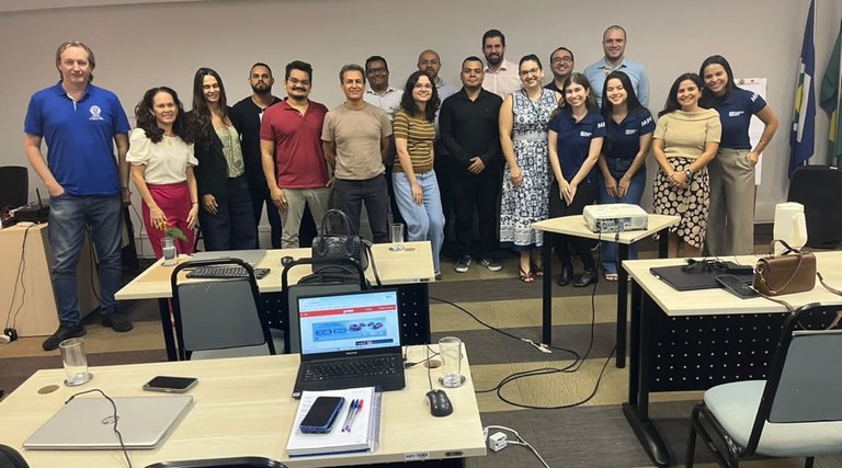 Em um sala com computadores sobre mesas e outros materiais de expediente, um grupo de pessoas está de pé posando para foto