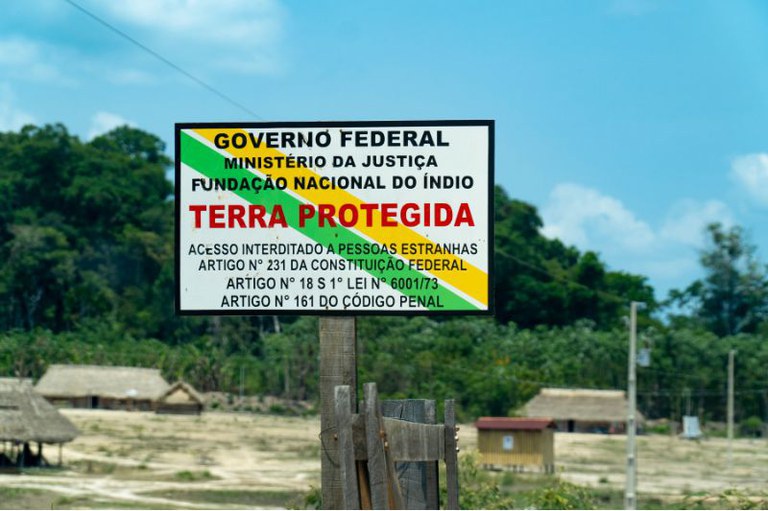 Placa oficial da Funai e do Governo Federal com os dizeres "Terra Protegida - Acesso interditado a pessoas estranhas", em primeiro plano, com habitações indígenas e floresta ao fundo.